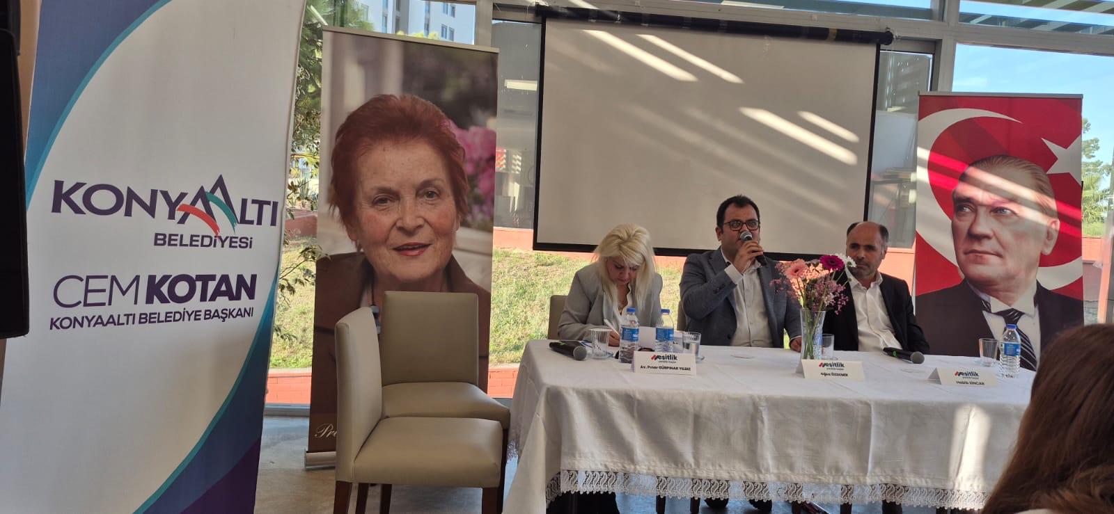 Bir belediye etkinliği/panel sahnesi.  Kapalı ama camlı, gün ışığı alan bir salonda uzun bir masa var; masa beyaz örtülü. Masanın arkasında sol başta Avukat Pınar GÜRPINAR YILDIZ, ortada Genel Başkanımız Oğuz ÖZDEMİR ile en sağda Genel Sekreterimiz Habib SİNCAR  oturuyor. Genel Başkanımız Oğuz ÖZDEMİR elinde mikrofonla konuşuyor, Avukat Pınar GÜRPINAR YILDIZ önündeki kâğıtlara eğilmiş, not alıyor, Genel Sekreterimiz Habib SİNCAR ise dinliyor. Masanın üzerinde su şişeleri, bardaklar ve ortada pembe çiçekli bir vazo, masanın önünde yerde iki el mikrofonu görünüyor.  Arka planda büyük bir projeksiyon/perde, Sağ tarafta Türk bayrağı fonu önünde Mustafa Kemal Atatürk’ün büyük bir portresi bulunan bir pano/dikey afiş duruyor. Sol tarafta ise “Konyaaltı Belediyesi” yazılı bir roll-up ve yanında yaşlı bir kadının büyük portresinin bulunduğu başka bir afiş bulunuyor.  Solda roll-up üzerindeki görünen metin: KONYAALTI BELEDİYESİ CEM KOTAN KONYAALTI Belediye Başkanı yazıyor.